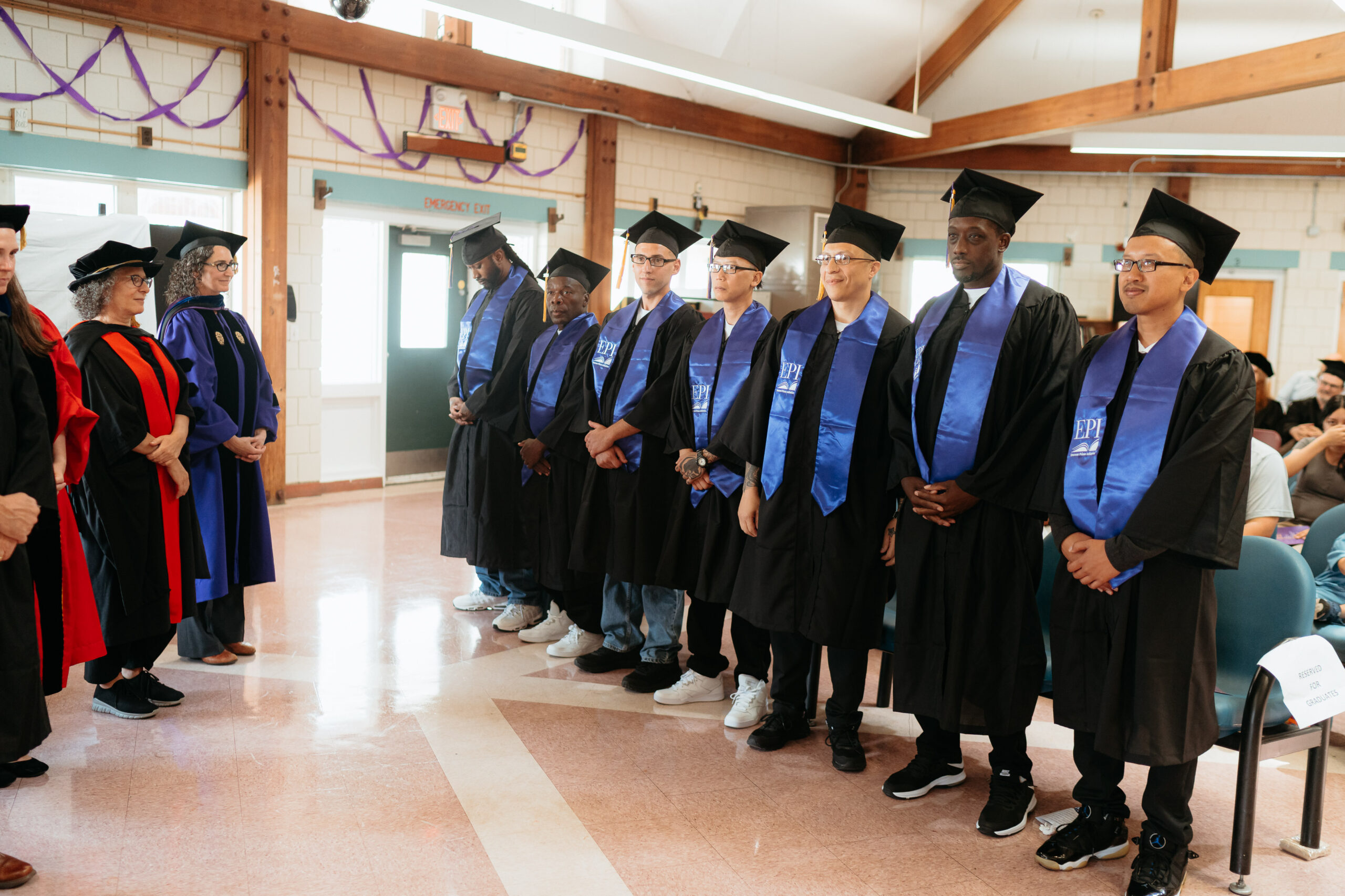 EPI graduates standing in a row, wearing regalia, facing College leadership
